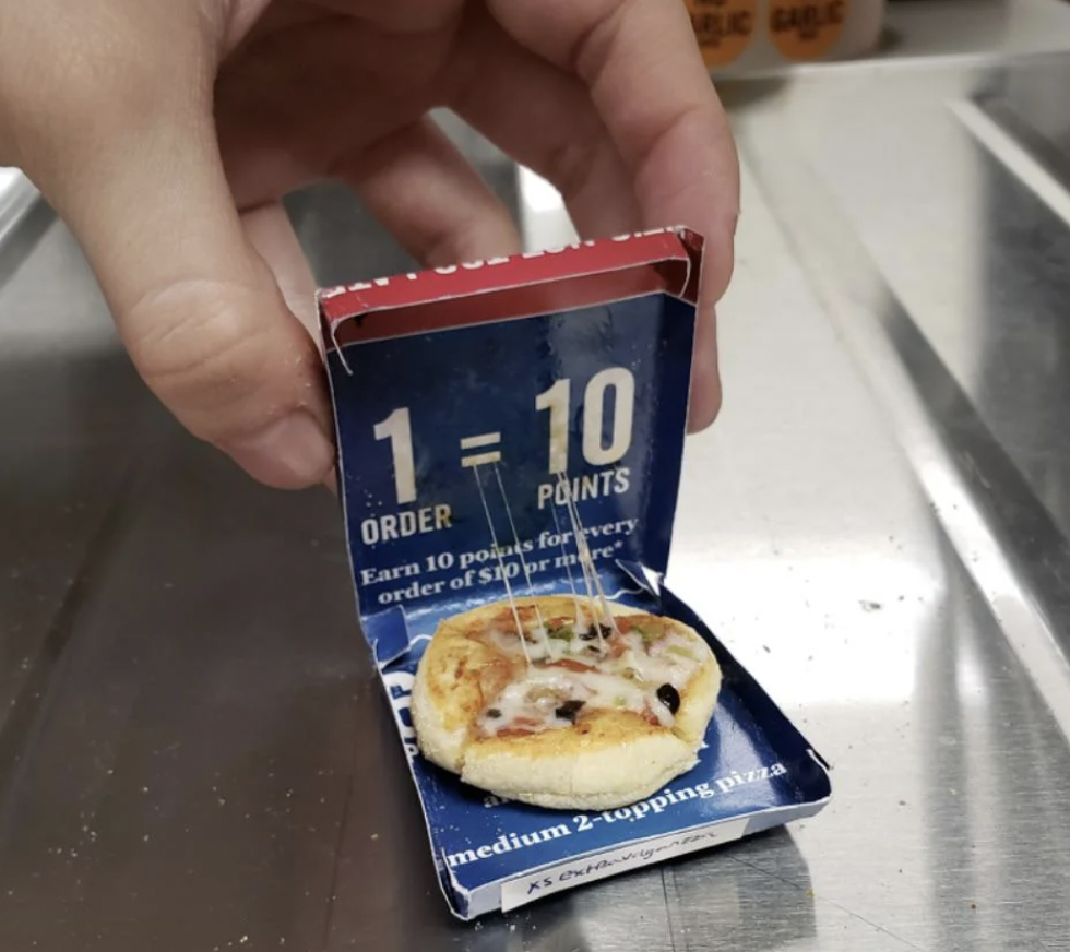 A hand holds a tiny pizza box with an equally small pizza inside it. The box has text stating "1 ORDER = 10 POINTS. Earn 10 points for every order of $10 or more." The pizza has melted cheese and visible toppings. The scene is set on a stainless steel surface.