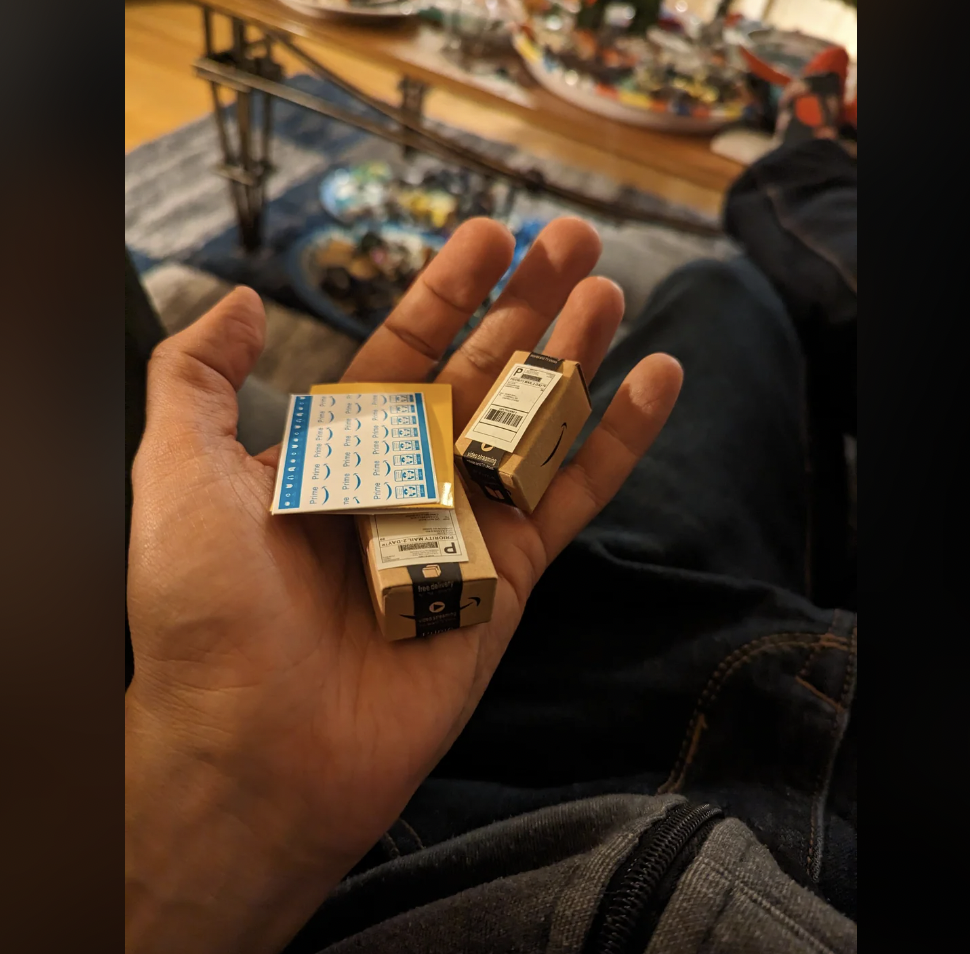 A hand holding two ink cartridges and a sheet of small sticker labels. The background shows a blurred view of a room with scattered items and a partially visible person sitting on a couch.