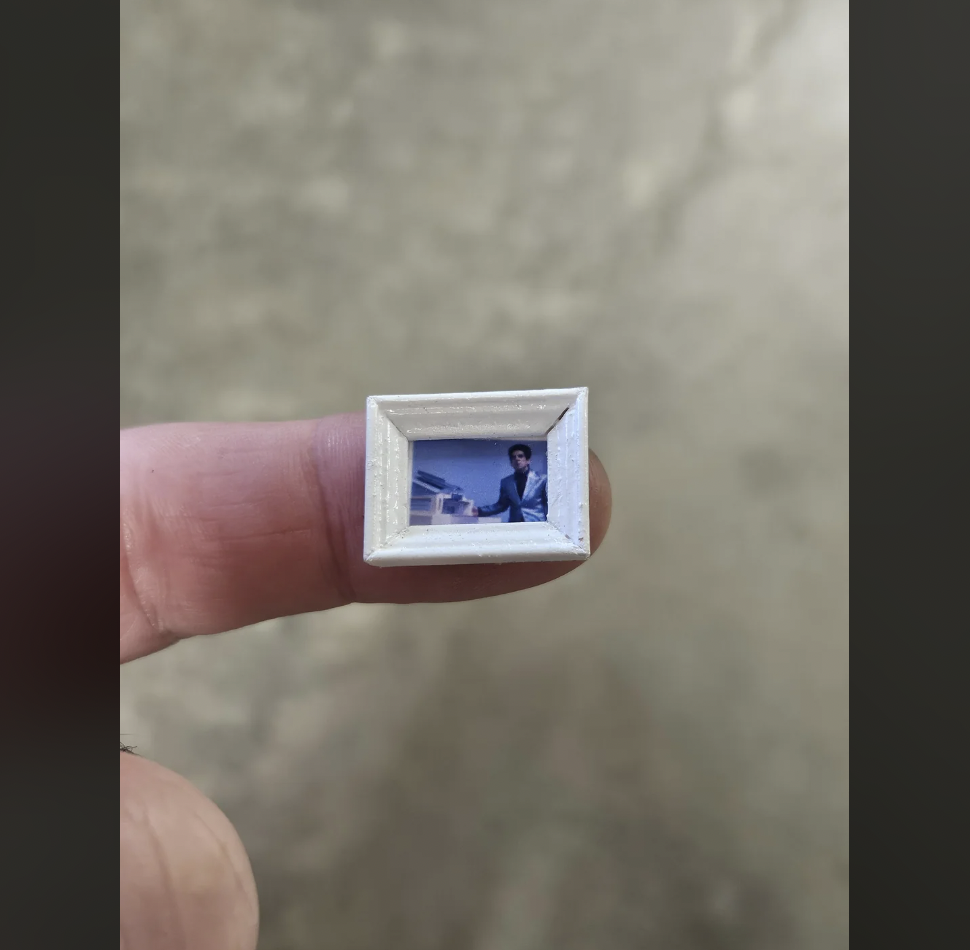 A person's fingers hold a tiny framed picture depicting a man sitting at a desk, with what looks like a building in the background. The detailed miniature photograph contrasts with the rough texture of the fingers and the blurred neutral background.