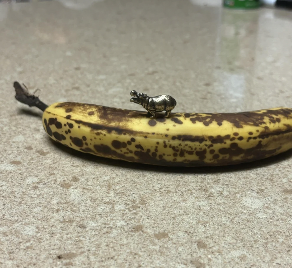 A small, intricately detailed figurine of a rhinoceros placed on top of a ripe, brown-spotted banana, which rests on a beige countertop.