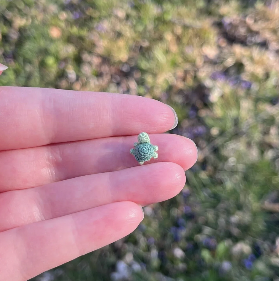 A close-up of a hand holding a tiny green crocheted turtle between the thumb and index finger. The background is a blurred outdoor scene with green grass and scattered purple flowers.