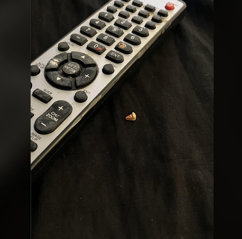 A TV remote control lies on a black fabric surface next to a small, triangular orange snack crumb. The remote has multiple buttons, including numbers and directional arrows, and an orange power button at the top.