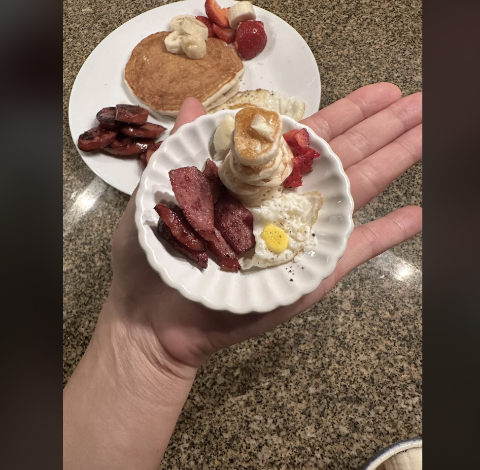 A hand holds a miniature plate with tiny pancakes topped with butter, small slices of strawberry, a tiny fried egg, and small pieces of sausage. A regular-sized breakfast plate with similar items is visible in the background on a granite countertop.