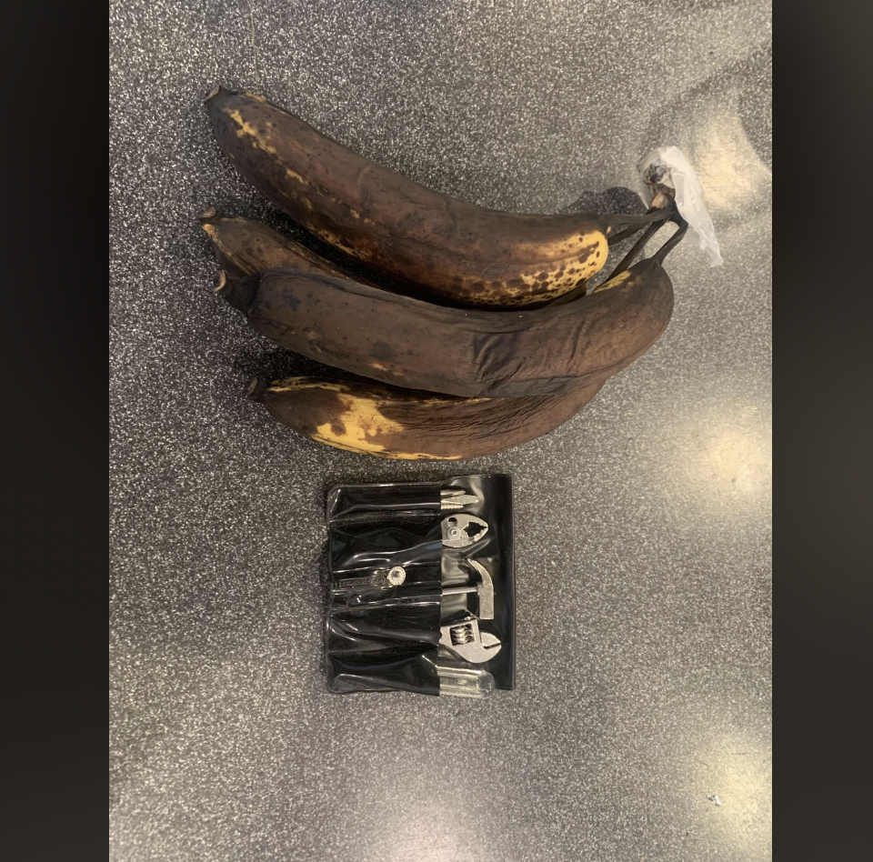 A bunch of overripe bananas with brown and black spots is placed on a gray countertop. In front of it is a black plastic packaging containing six small metal bottle openers with black handles.