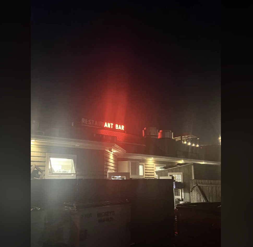 A restaurant is dimly lit at night, featuring a red neon sign that reads "RESTAURANT BAR." The building appears to be wooden with lights on inside. There is a fenced area and some dumpsters are visible in the foreground. The surrounding area is dark.