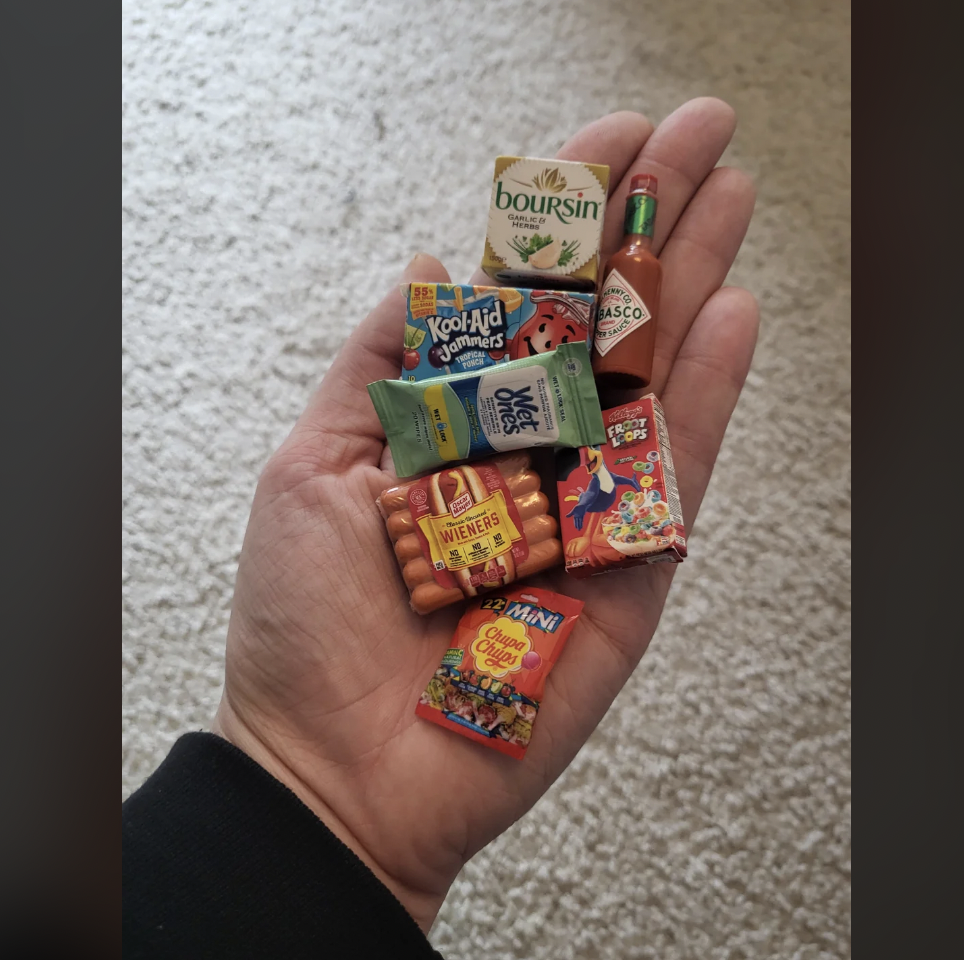 A hand holds miniature-sized items including packages of Boursin cheese, Kool-Aid Jammers, Rice Krispies Treats, Tabasco sauce, wieners, Colossal Crunch cereal, and What the Frittle. The background is a carpeted floor.