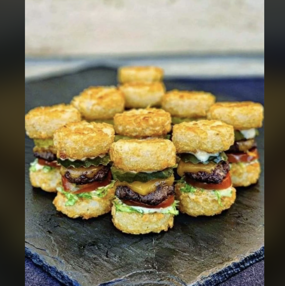 A plate of small sliders made with tater tots as buns. Each slider consists of a piece of meat, melted cheese, a pickle slice, lettuce, and a slice of tomato, stacked between two tater tots, all arranged on a slate serving board.