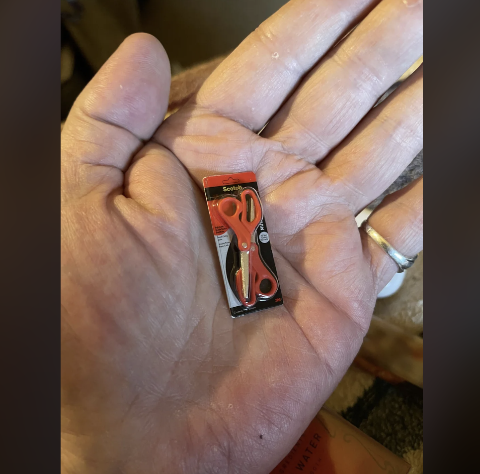 A close-up of a hand holding a tiny, toy-sized pair of red scissors in its packaging. The packaging is black and red, labeled "Scotch." The hand wears a silver ring on the ring finger.