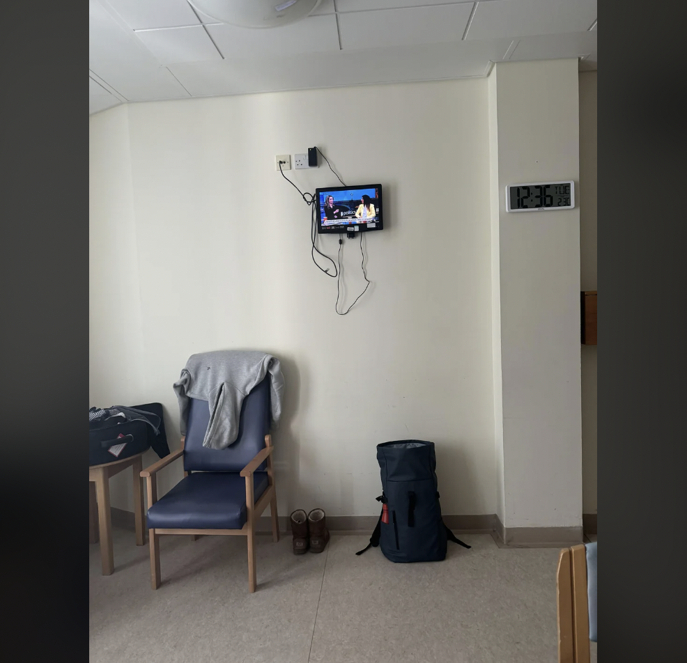 A small wall-mounted TV showing a news program in a room with a blue chair covered by a gray blanket. Nearby are a pair of shoes, a large blue backpack, a wall clock showing 12:36, and some cables on the pale wall.