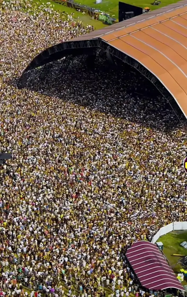 Aerial view of a massive crowd packed together tightly in front of a large stage with an arched roof, suggesting a large-scale concert or event. The sea of attendees appears dense, extending back as far as the eye can see.