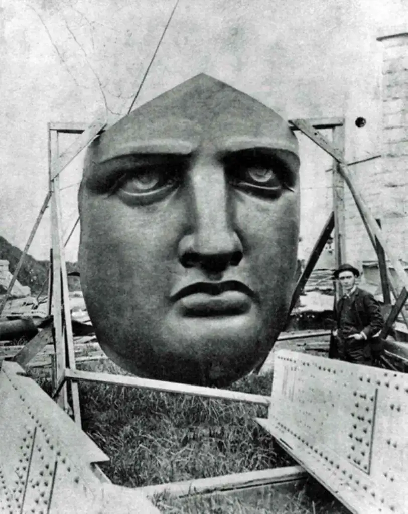 A historical black-and-white photo shows a man standing next to the large, detached face of the Statue of Liberty while it is under construction. The massive face is supported by a wooden frame at an outdoor workshop.