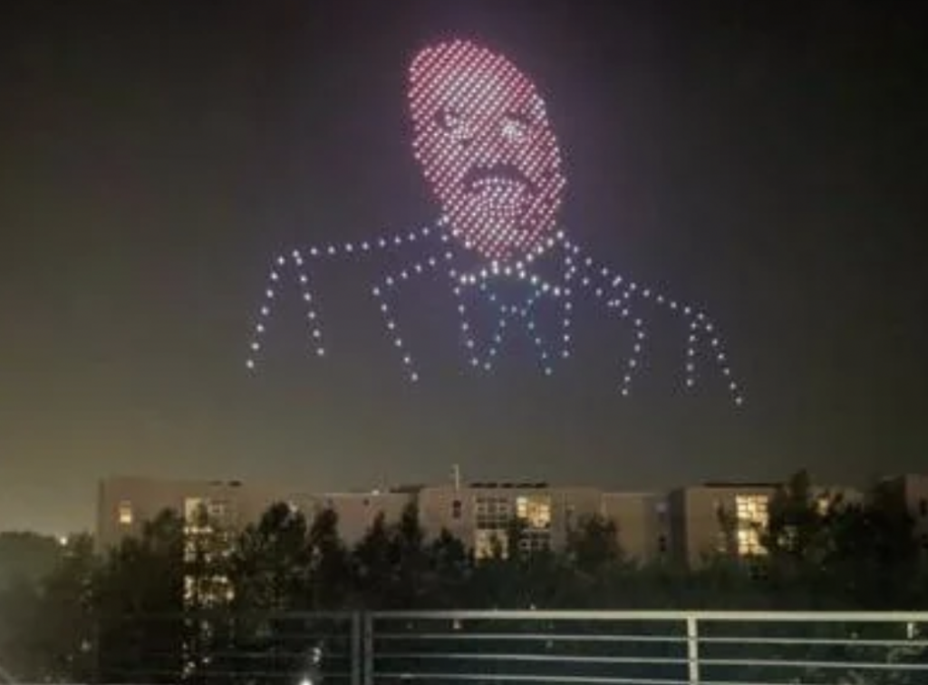 A night sky displays a drone light show forming the image of a man in a suit and bow tie. The vibrant lights create detailed features of the man's face and clothing. Trees and a row of buildings are visible in the background at the bottom of the image.