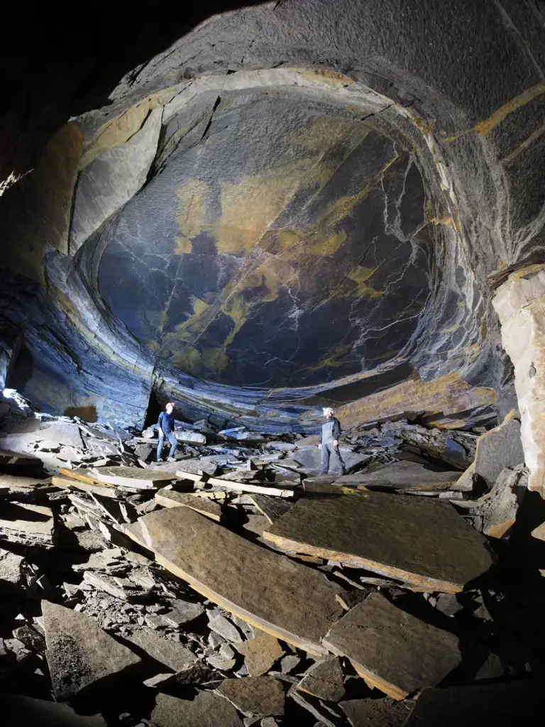 Two explorers stand inside a large, abandoned slate quarry cavern. The cavern features a huge, circular, dome-like ceiling with varied rock textures and hues. Broken slate pieces are scattered across the ground, and the explorers are illuminated by their headlamps.