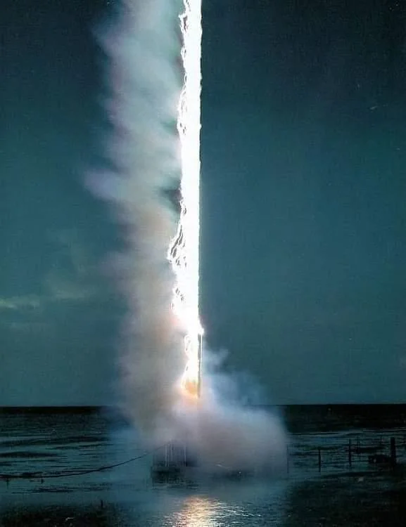 A bright lightning bolt strikes a body of water at night. The bolt creates a vivid, vertical line of light, surrounded by mist and vapor rising from the impact site. The surrounding area is dark, with an indistinct horizon and a cloudy sky.