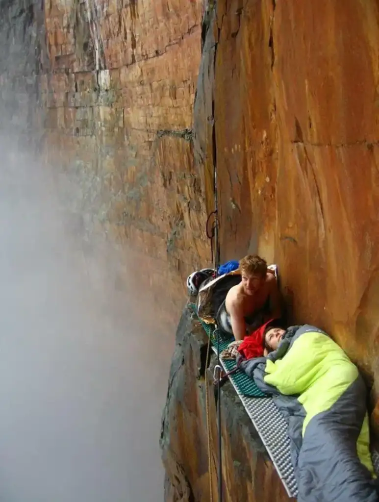 Two climbers rest on a narrow, suspended platform halfway up a steep, orange-hued cliff face. One climber is wrapped in a green sleeping bag while the other sits shirtless beside them, against a backdrop of mist and rock. Climbing gear is visible around them.