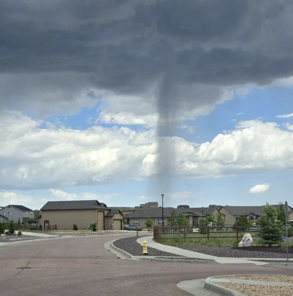 A narrow tornado or funnel cloud descends from a dark, cloudy sky towards a suburban neighborhood. Houses and a quiet street are visible in the foreground, with a yellow fire hydrant near the center of the image. Trees and shrubs dot the landscape.