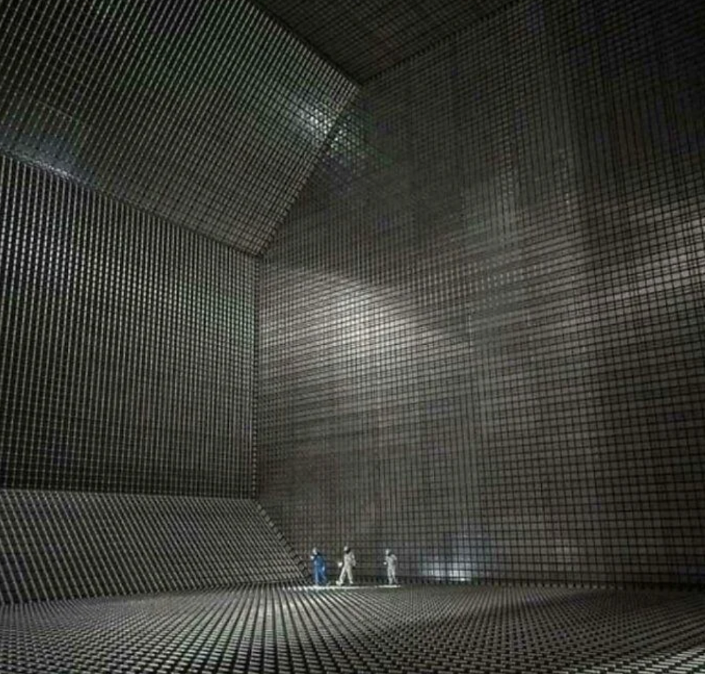 Three small figures in protective suits stand inside a cavernous, grid-lined metallic chamber with towering walls and a high ceiling, giving a sense of massive scale and industrial or futuristic environment.