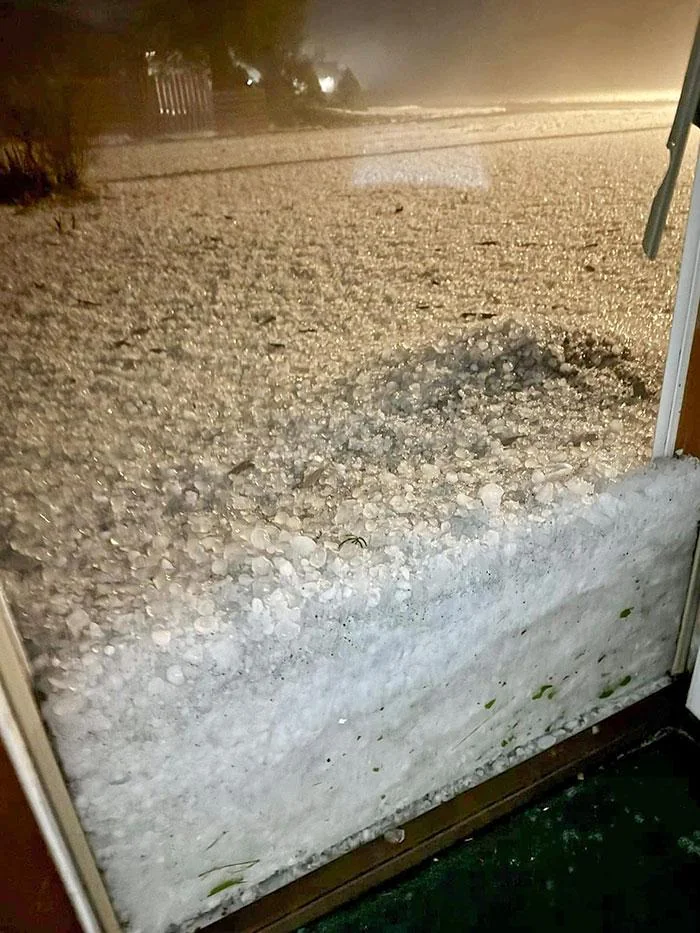 A view through an open door showing a thick accumulation of hailstones covering the ground and partially blocking the entrance. The scene outside is misty, creating an obscured atmosphere, with hail piled up against the door and spread out over the yard.