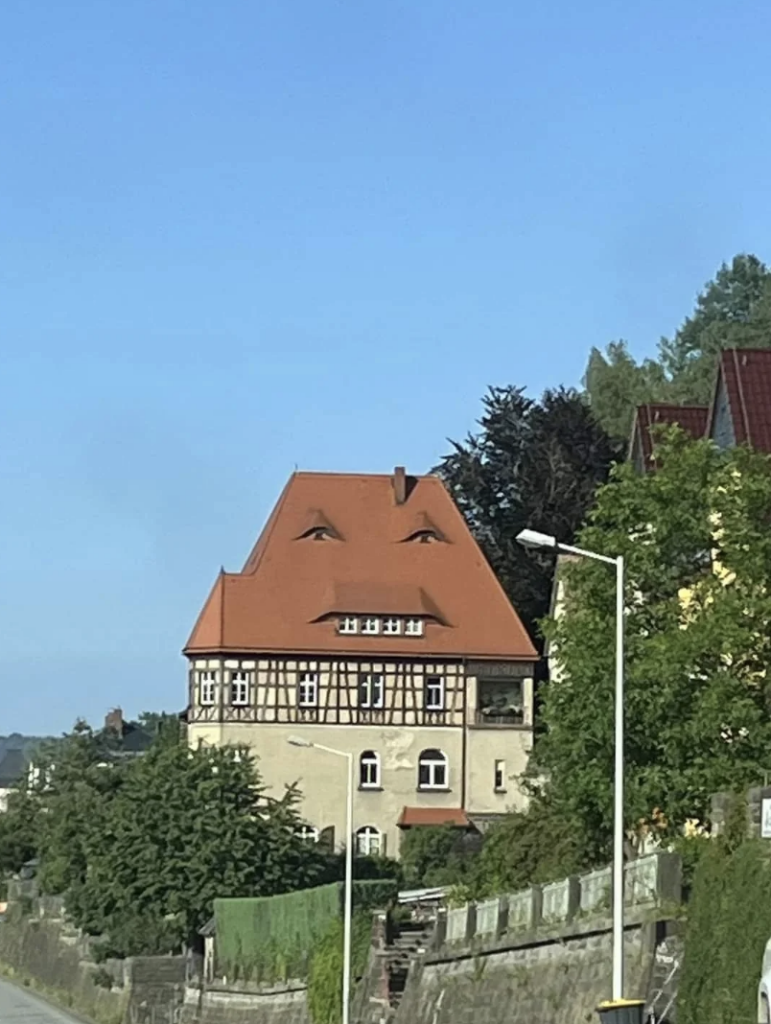 A building with a triangular, orange-red roof appears to have a face. Two windows resembling eyes are positioned near the top, and a small, central window below resembles a nose. The rooflines and cornices suggest a smiling mouth. The sky is clear and blue.