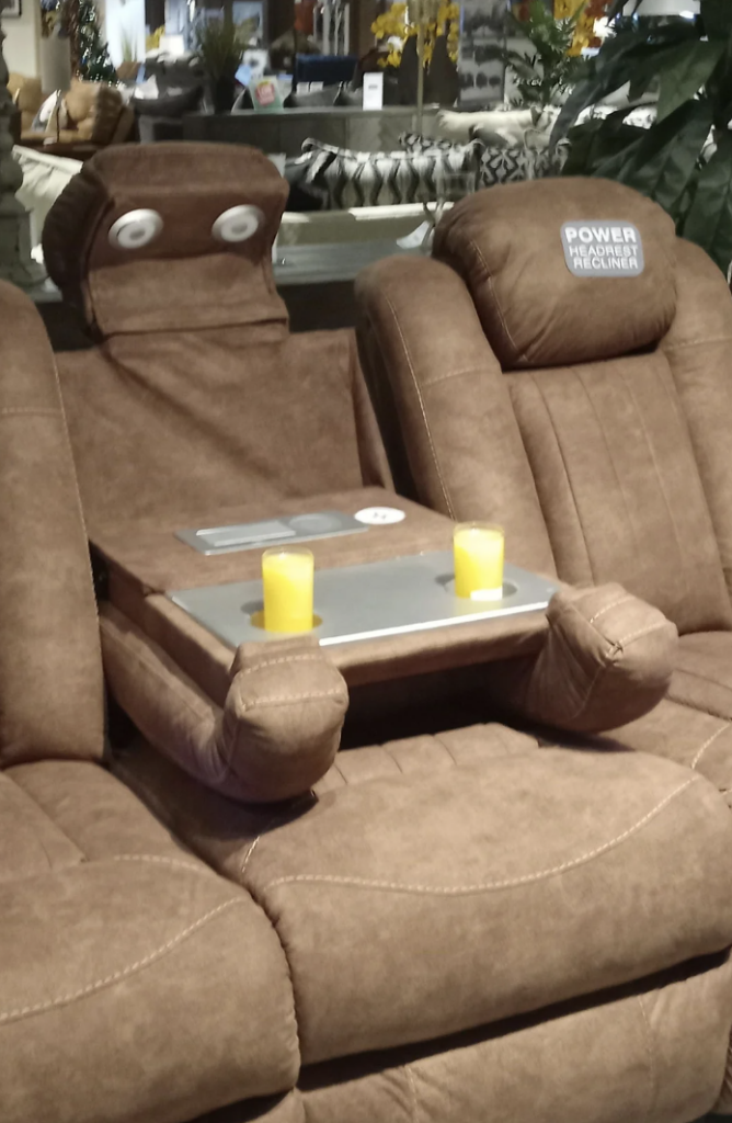 A brown reclining sofa with a unique design where the middle seat appears to resemble a face, with two round buttons as eyes. The middle backrest is lowered to act as a tray, holding two yellow candles. The sofa has a "Power Recliner" label.