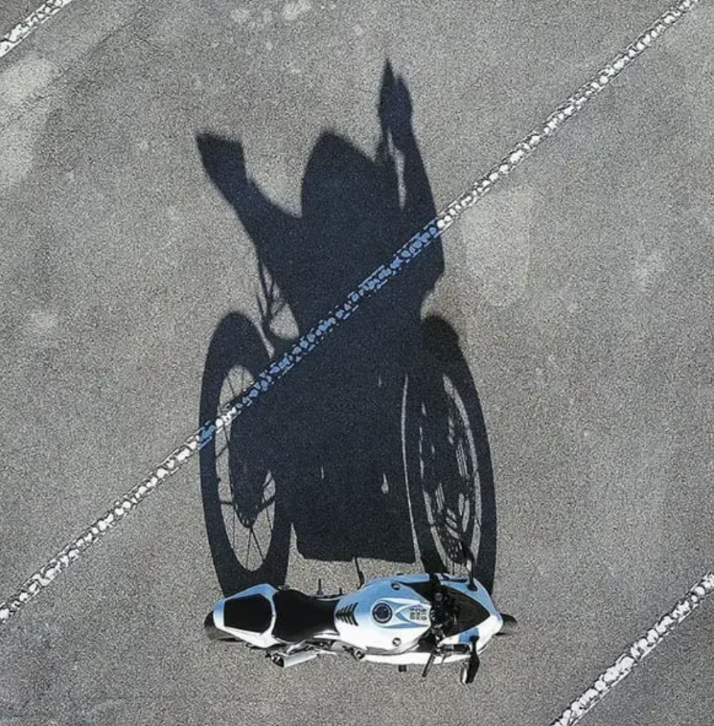 Aerial view of a motorcycle parked on an asphalt surface, casting a shadow that visually resembles a person with arms raised, creating an illusion of a rider with exaggerated gestures due to the angle of the sunlight.