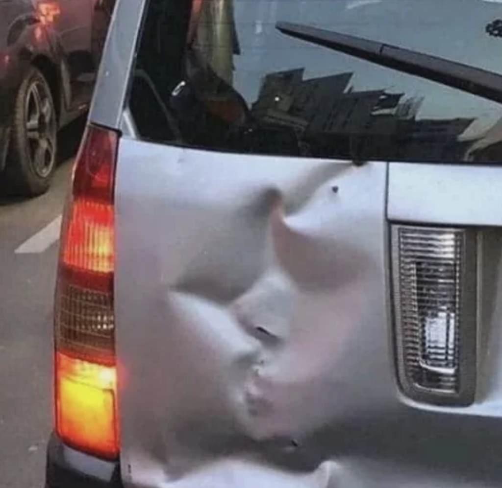 A close-up image of the back of a silver car with a noticeable dent on its rear door. The dent is located near the right tail light, which is illuminated. Another vehicle is visible in the background.