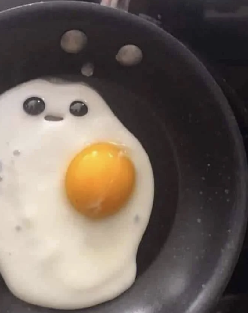 A fried egg in a pan appears to resemble a ghost face with eyes and a mouth. The egg yolk serves as the ghost's body and the shape of the egg white forms its head.