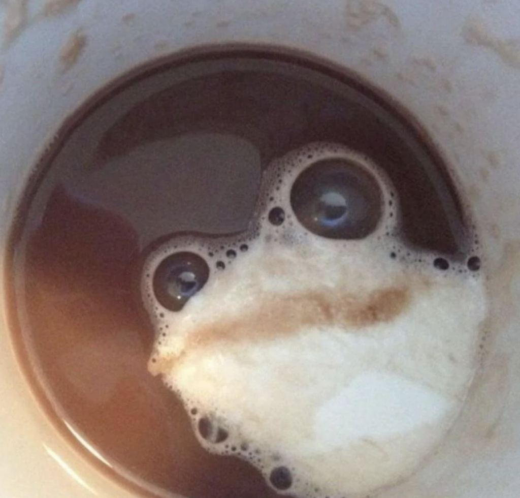 A close-up of coffee with cream forming a pattern resembling a surprised face. The face has two large bubbles as eyes and a mustache-like creamy layer below them, creating an amusing, anthropomorphic impression.