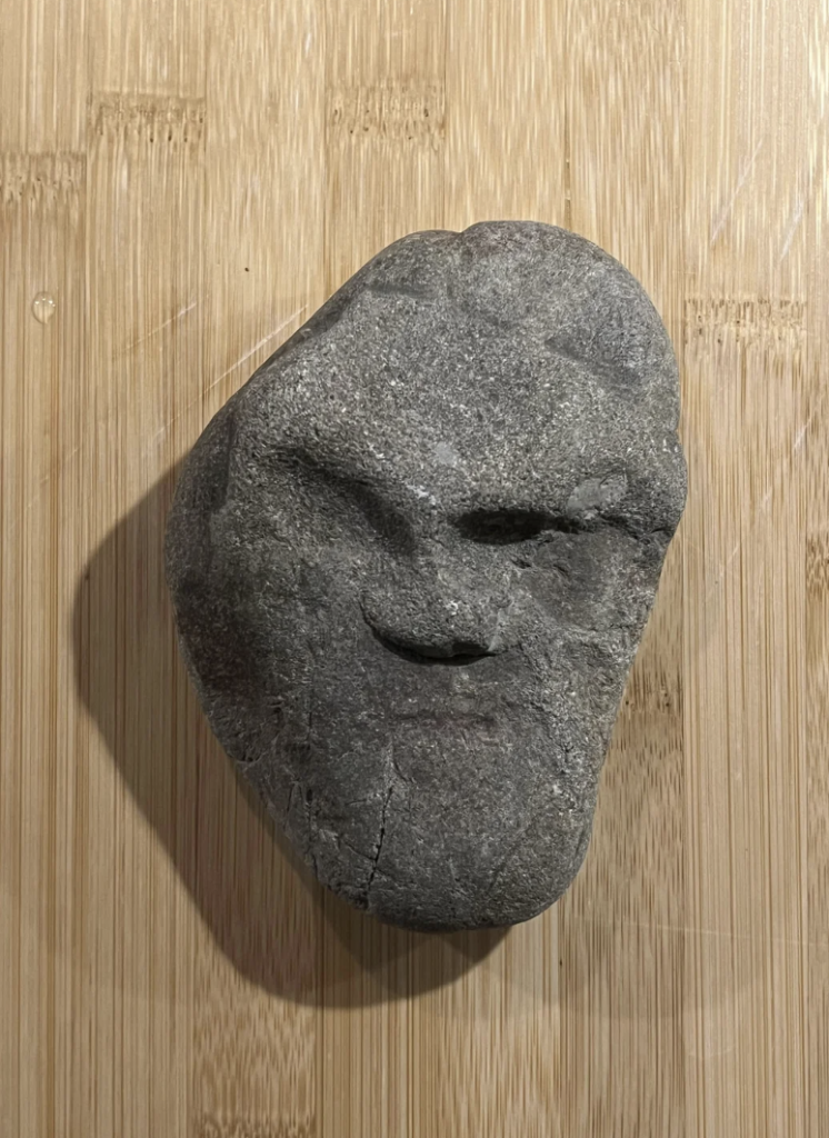 A gray rock resembling a human face with carved-like features lies on a light wooden surface. The rock's natural contours give the appearance of eyes, a nose, and a mouth.