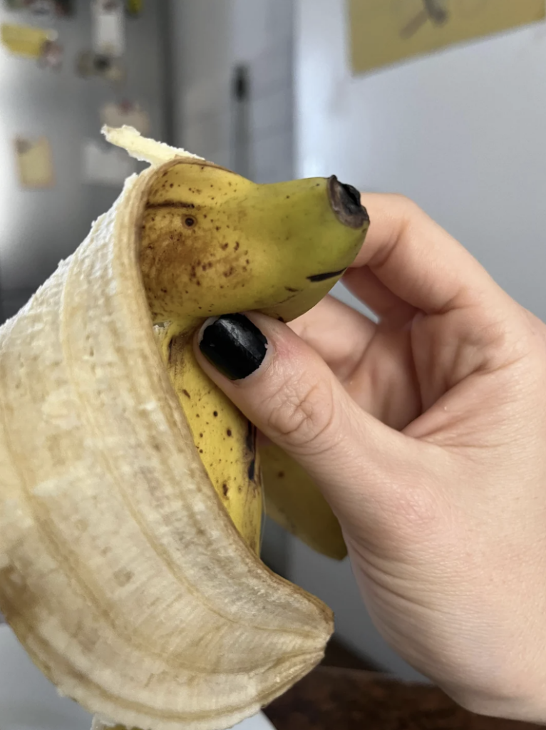 A peeled banana being held up by a hand with black painted nails. The top part of the banana, which is not peeled, has been creatively shaped and drawn to resemble a dog's face. The banana peel is pulled to the sides, resembling the dog's ears.