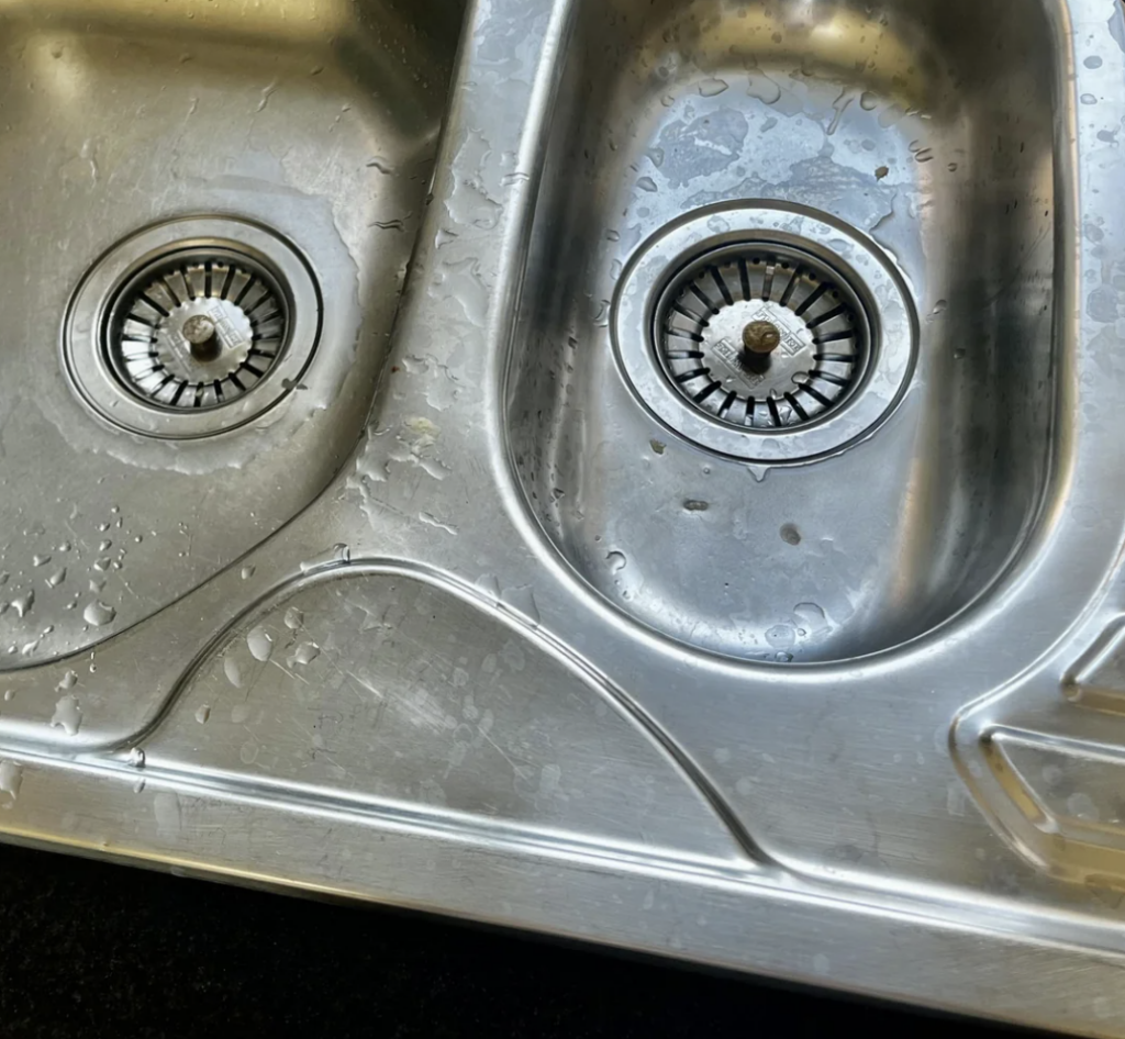 A stainless steel double kitchen sink with water droplets visible. Both basins have circular drain covers with one on the left and the other on the right. The sink surface shows slight wear with scratch marks.