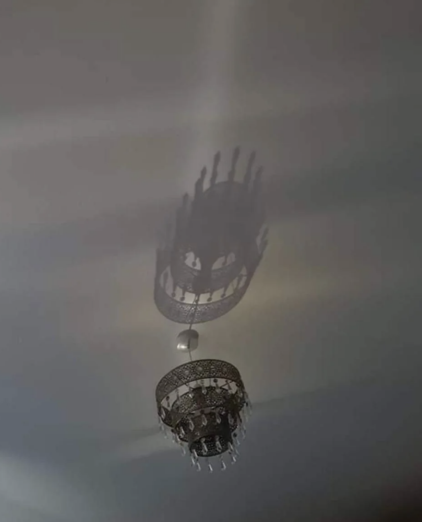 A decorative chandelier is attached to a ceiling, casting a detailed shadow on the surface above. The chandelier has intricate patterns and hanging ornaments, creating an elegant ambiance with its shadowy reflection.