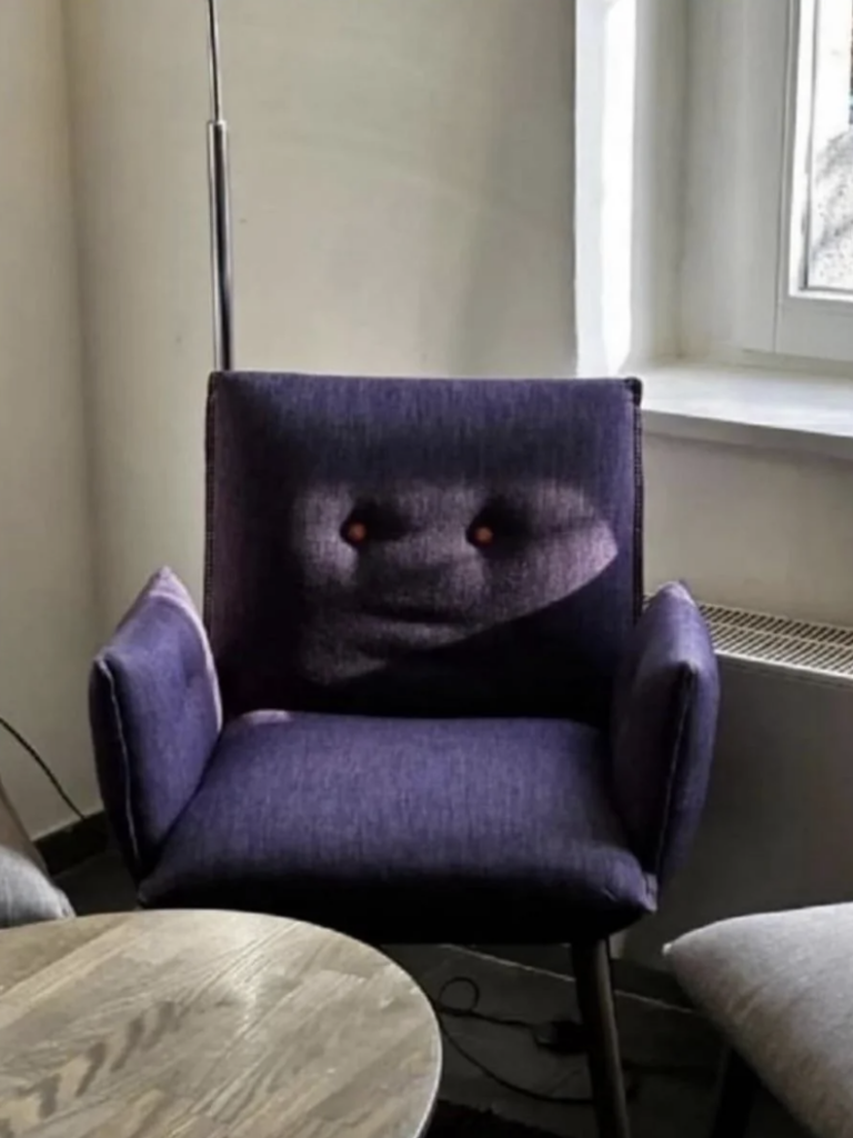 A purple cushioned chair with armrests sits in a corner next to a window. The cushion's wrinkles and shadows create an illusion of a face with button-like eyes, giving the chair a whimsical, expressive appearance. A round wooden table is partially visible in the foreground.