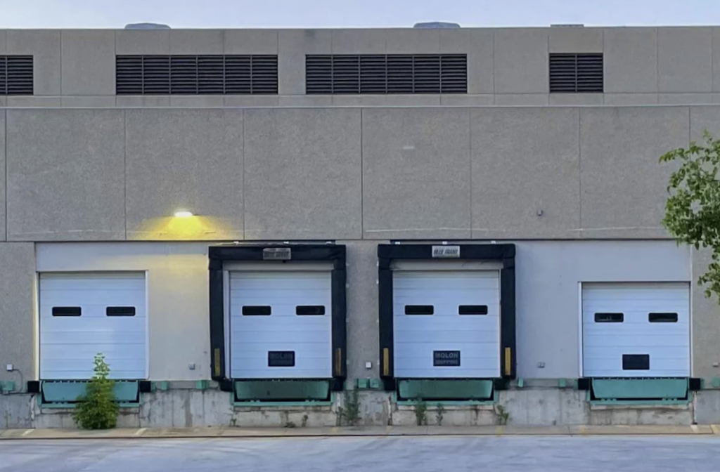 A concrete building with three loading dock doors, each equipped with a metal dock leveler and rubber bumpers. The center door has a "Section 7" label. A small tree is visible on the right side, and a light above the left door illuminates the area.