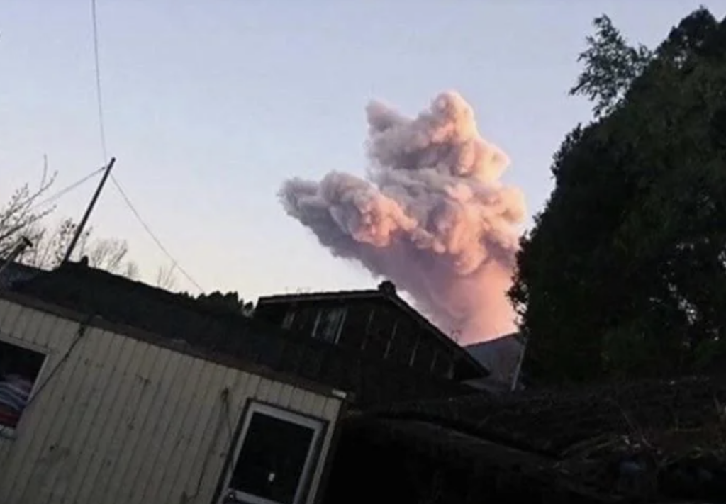 A large plume of ash and smoke rises into the sky from a distant volcano, with the foreground showing an angled view of nearby buildings and trees. The eruption is highlighted by the soft pink and orange hues of the setting or rising sun.