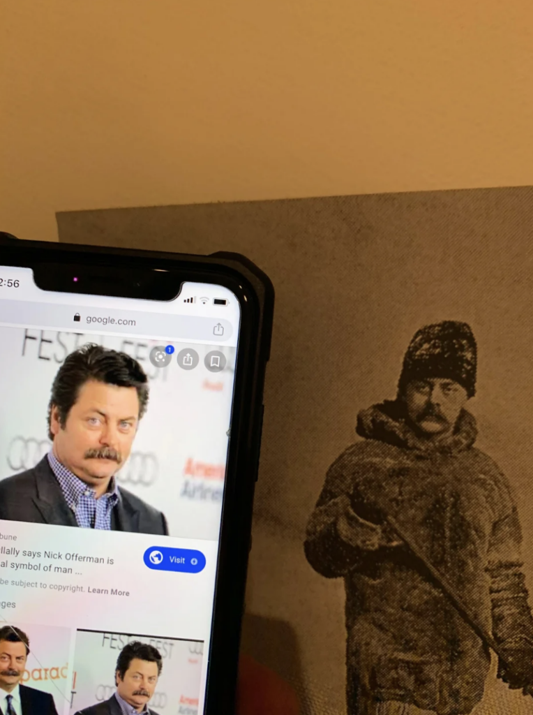 A smartphone displays an image of a man with brown hair, a mustache, and a blue shirt. Next to the phone, there is an old black-and-white photo of a man with a similar mustache, wearing winter gear and holding a rifle.