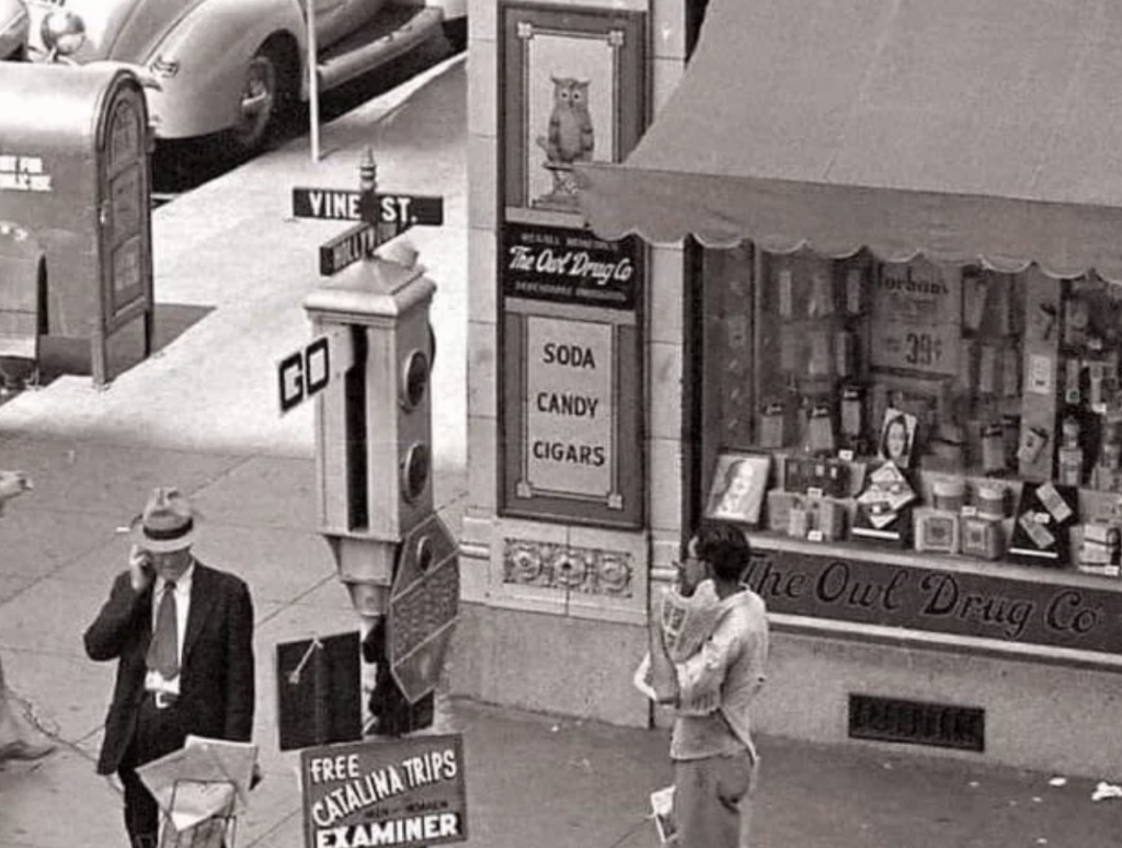 A black-and-white photo shows a street corner with a drugstore named "The Owl Drug Co." The storefront displays various items. A man in a suit and hat uses a payphone, while a woman stands nearby holding a newspaper. There is an old traffic signal with the word "GO.