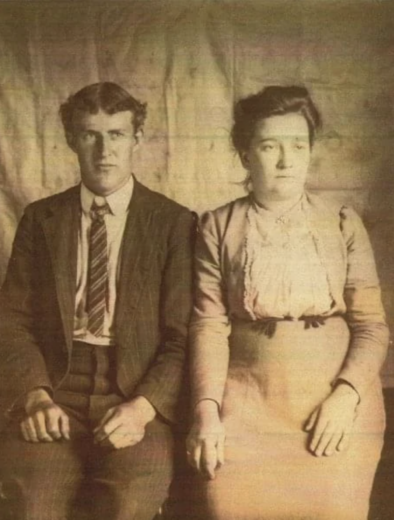 A sepia-toned vintage photograph of a man and a woman sitting side by side. The man, on the left, is wearing a suit and tie with a serious expression. The woman, on the right, is in a long-sleeved dress with lace details, also looking serious. The background is plain.