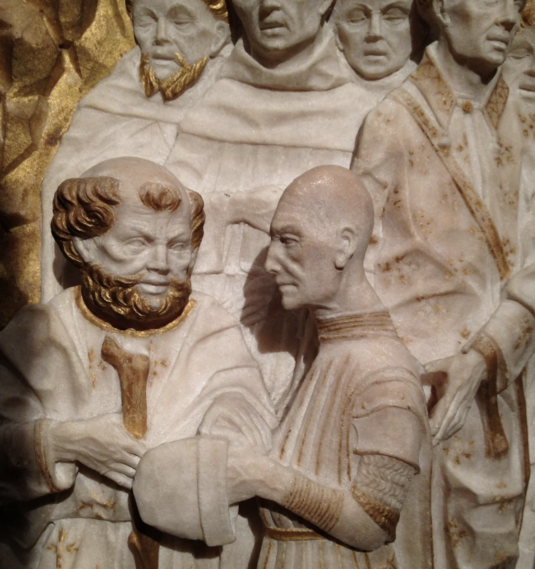 A detailed stone sculpture depicts two men in conversation. The man on the left has curly hair and a beard, holding a piece of fabric. The man on the right is bald with a craggy face. Other figures surround them, wearing robes and expressions of attentiveness.