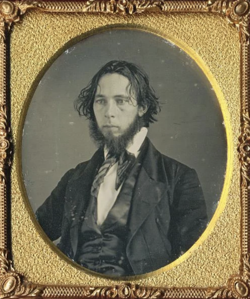 A daguerreotype photograph of a young man with long, dark hair and a distinctive beard. He is wearing a formal suit with a high-collared shirt, waistcoat, and a cravat. The photo is set in an ornate gold frame with intricate detailing.