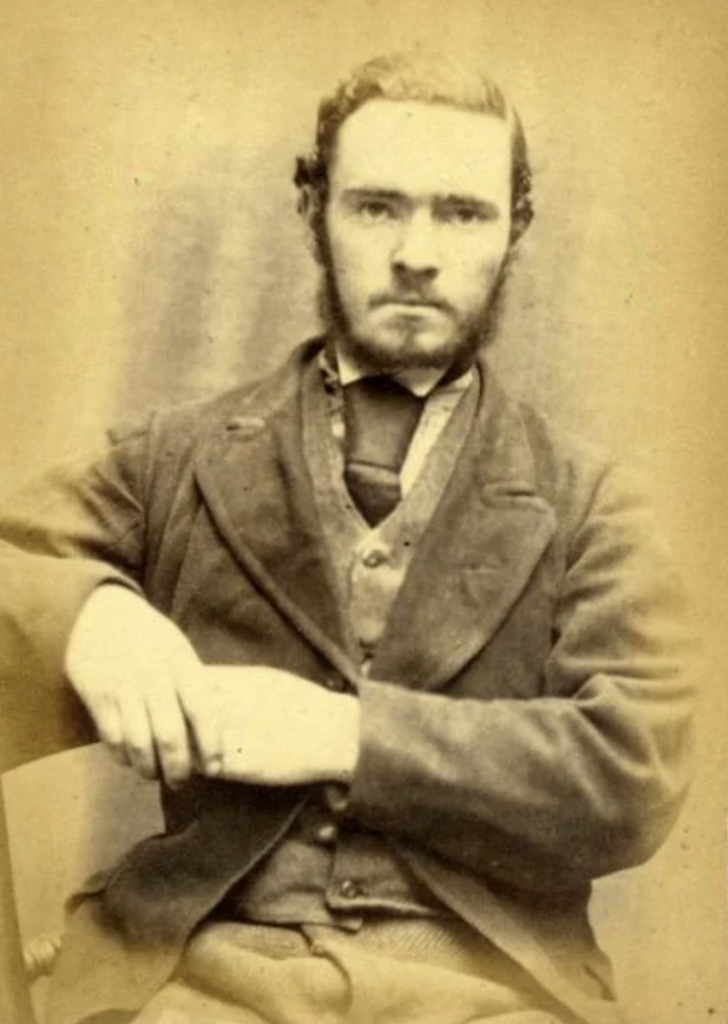 A sepia-toned portrait of a man from the 19th century. He is seated and wearing a formal suit with a vest, high-collared shirt, and tie. He has a serious expression, short hair, and a full beard with a mustache. His right arm rests across his lap.