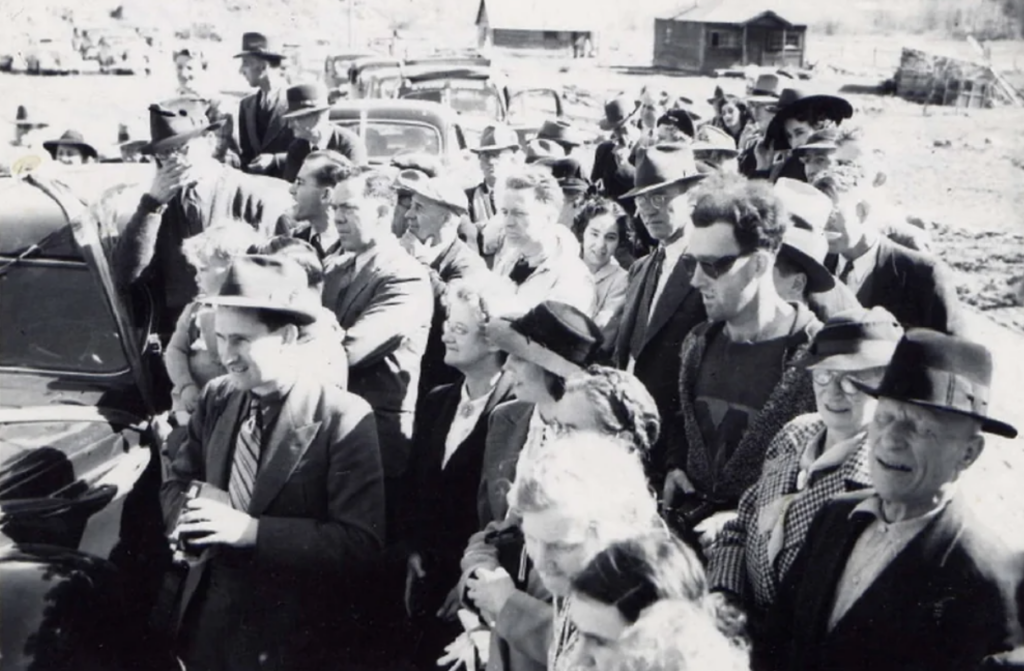 A black and white photo depicting a crowd of people, most wearing hats and coats, gathered outdoors next to a row of cars. In the background, there are rural buildings. The mood appears festive or communal. The photograph gives a vintage, mid-20th century feel.