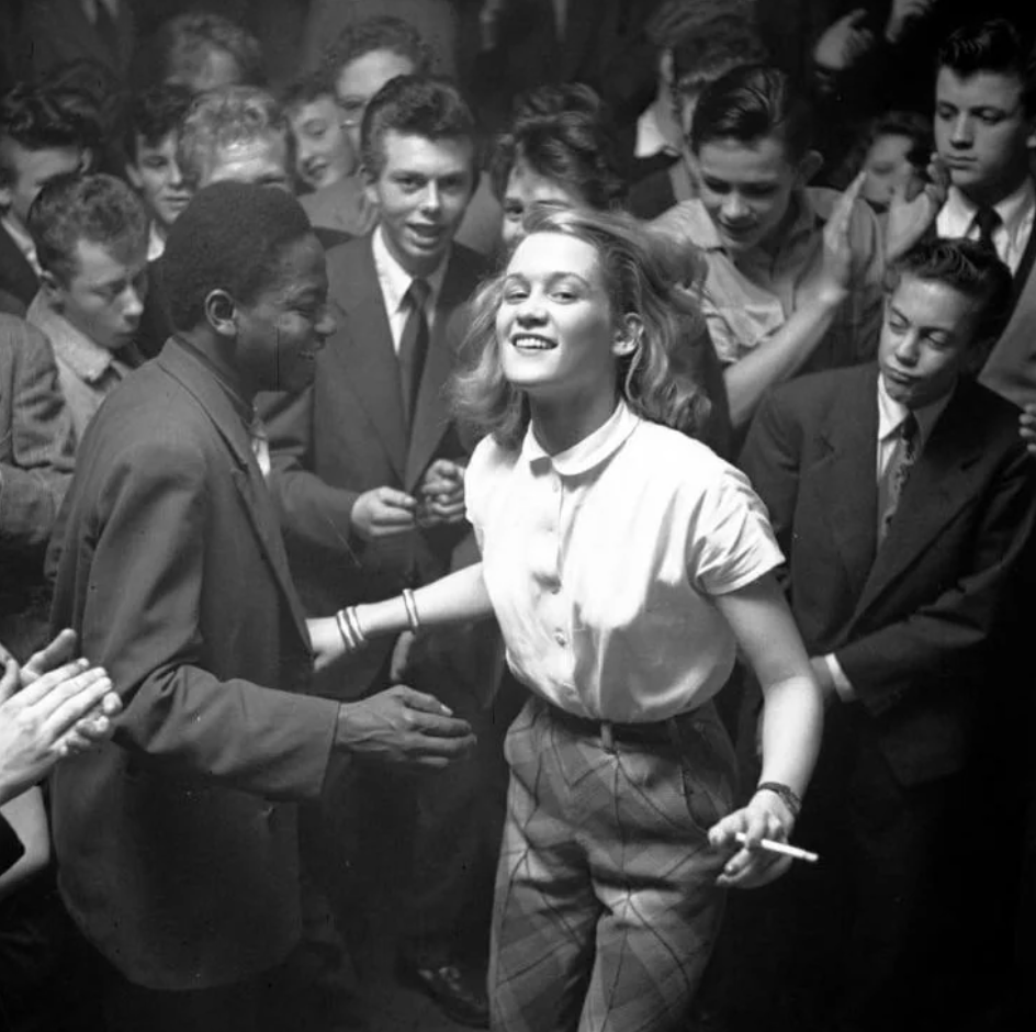 A black and white photo shows a lively dance scene from the 1950s or 60s. A woman and a man are dancing energetically in the center of a crowd of onlookers, who are clapping and cheering. The dancers and the crowd are dressed in vintage attire typical of the era.