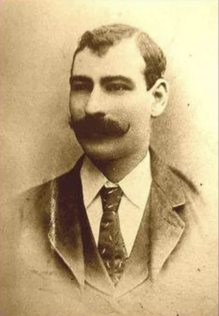 Sepia-toned portrait of a man with a thick mustache and short hair, wearing a suit with a tie and collared shirt. The background is plain, enhancing the vintage appearance of the image.