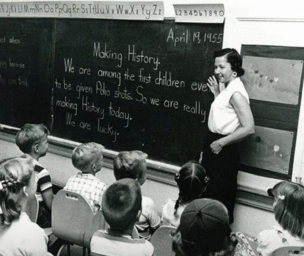 A teacher stands beside a chalkboard with children seated in front. The board reads, "Making History. We are among the first children ever to be given Polio Shots. So we are really making History today. We are lucky." The scene dates to April 19, 1955.