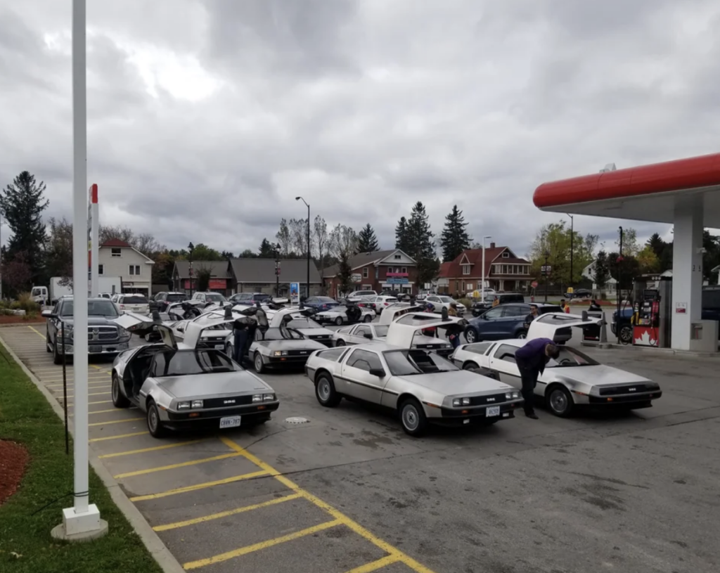 A group of DeLorean cars with gull-wing doors open are parked in rows at a gas station. Several people are seen around the cars, and some are working on them. The sky is overcast, and the surrounding area includes buildings and trees.