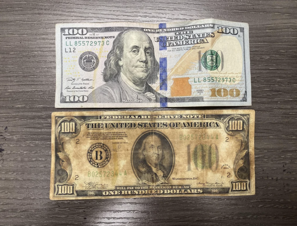 Two U.S. $100 bills are displayed side by side on a wooden surface. The top bill is a modern design with a large portrait of Benjamin Franklin and security features, while the bottom bill is an older design with a smaller portrait and a more prominent "The United States of America" text.