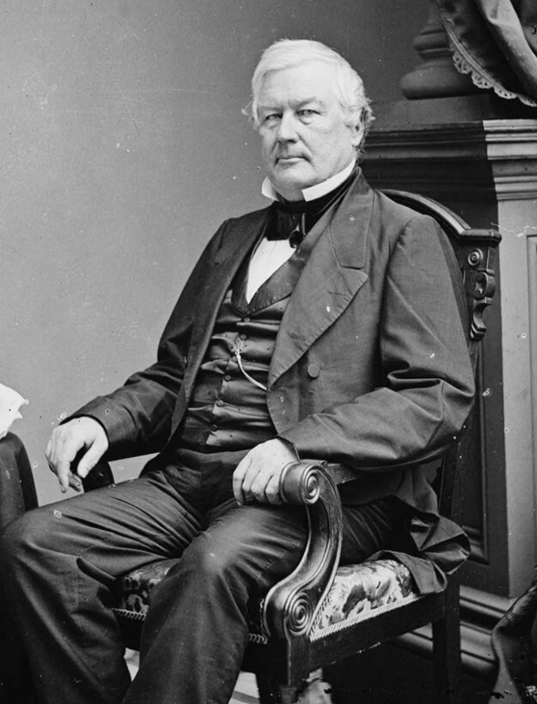 A black and white historical photograph of an older man seated in a formal wooden chair. He has white hair and sideburns and is wearing a dark suit with a vest, white shirt, and bow tie. The background features a draped curtain and part of a wooden cabinet or table.