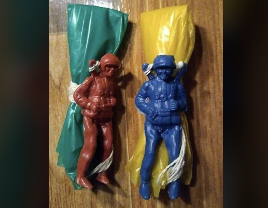 Two toy paratroopers are attached to wooden wall. One is red with a green parachute and the other is blue with a yellow parachute. Both have white cords connecting them to their packed parachutes.