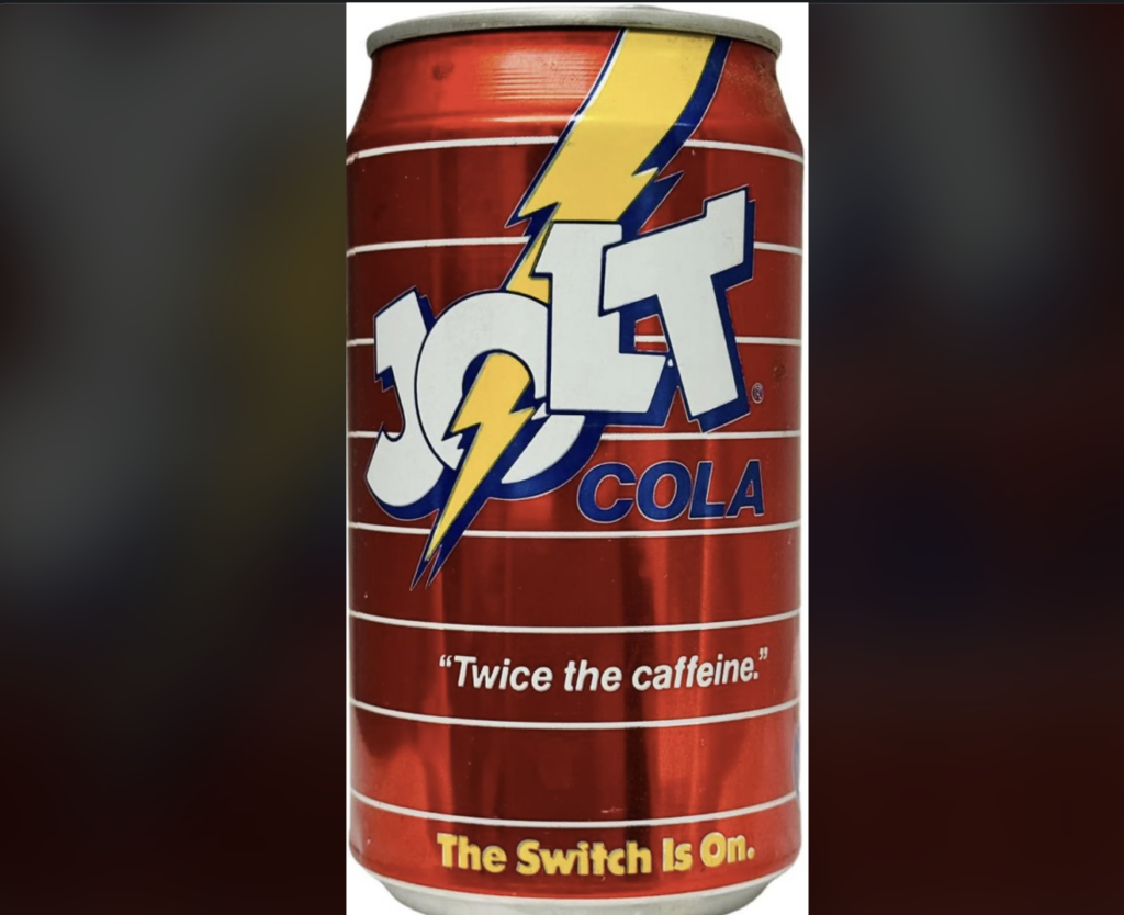 A red can of Jolt Cola featuring a yellow lightning bolt through the "O" in "Jolt." The can displays the slogan "Twice the caffeine" and "The Switch Is On" in yellow text at the bottom. The background has horizontal white lines.